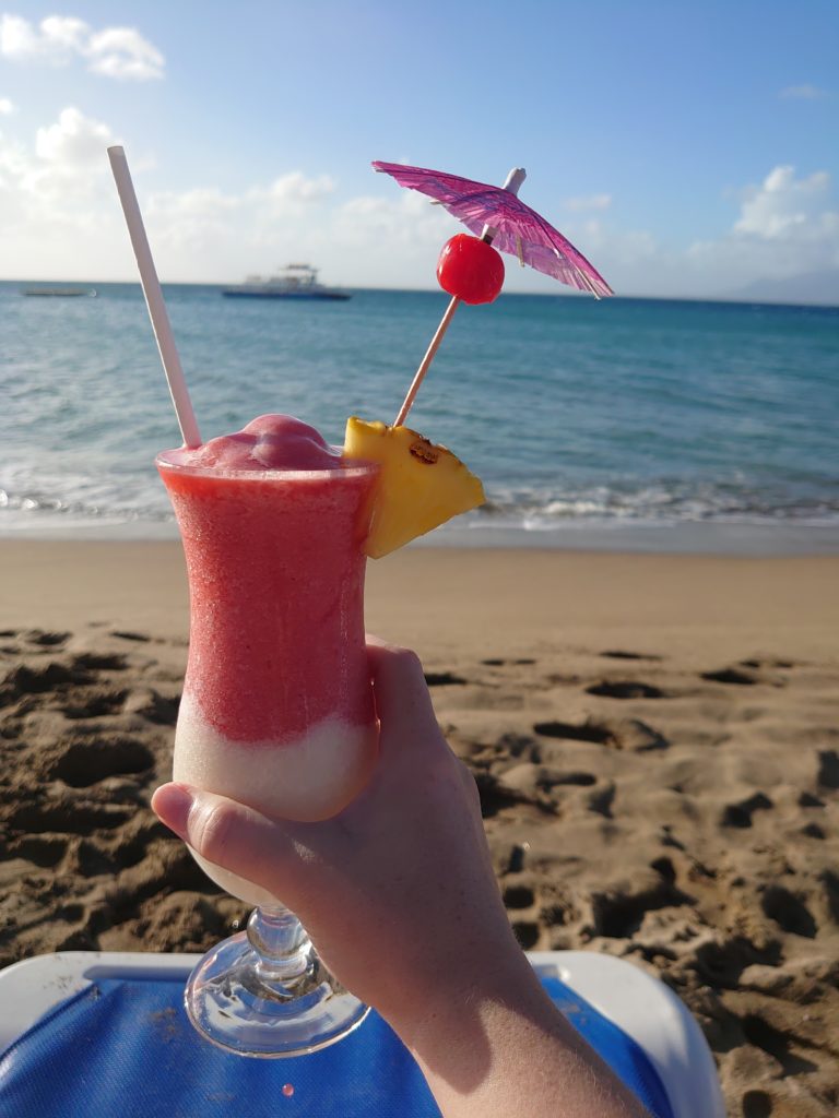 A hand holding a Miami Vice cocktail on a beach in front of the sea. One of the perks of living abroad