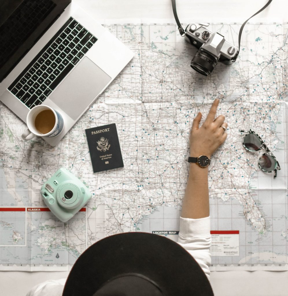 A person wearing a hat planning a trip abroad pointing at a map with a laptop, passport and camera