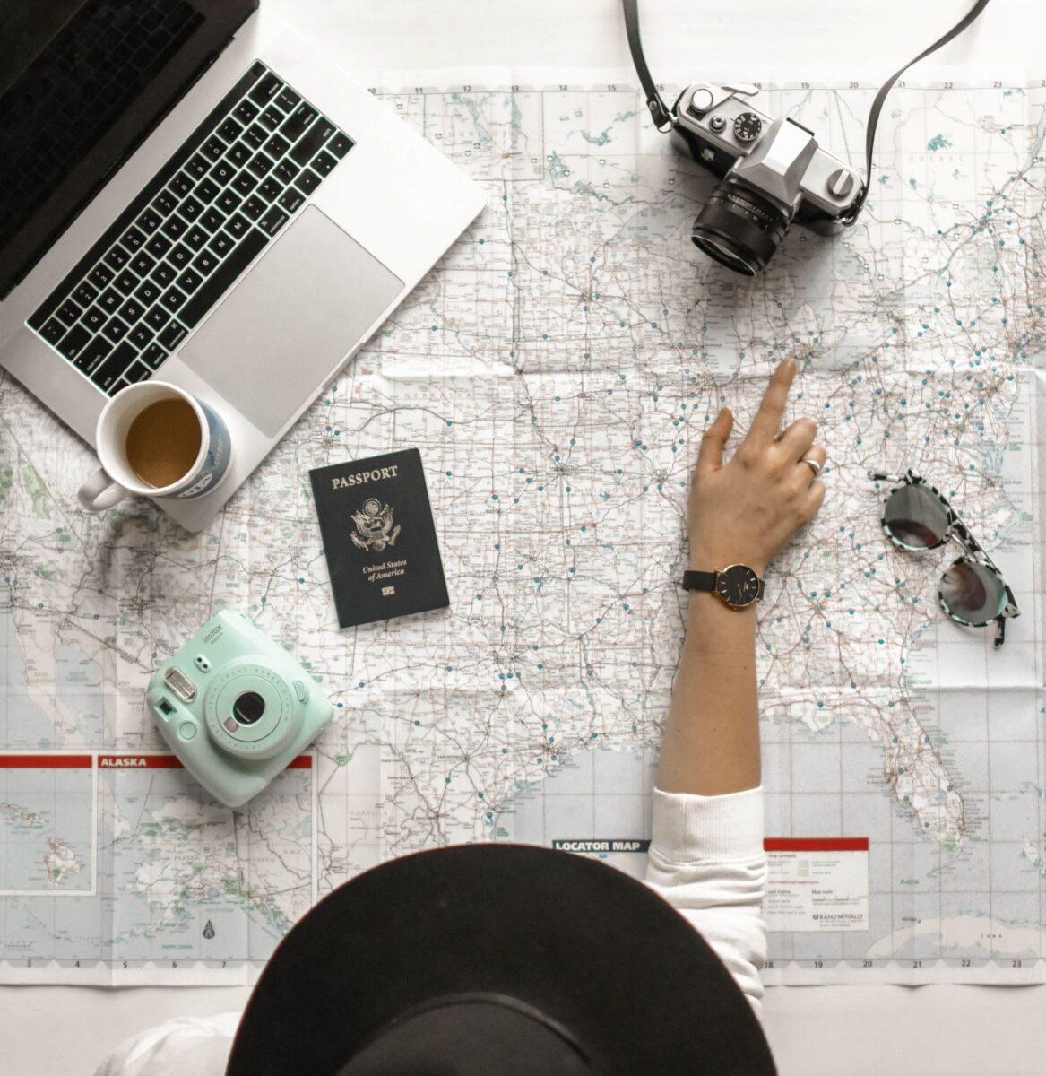 A person wearing a hat planning a trip abroad pointing at a map with a laptop, passport and camera