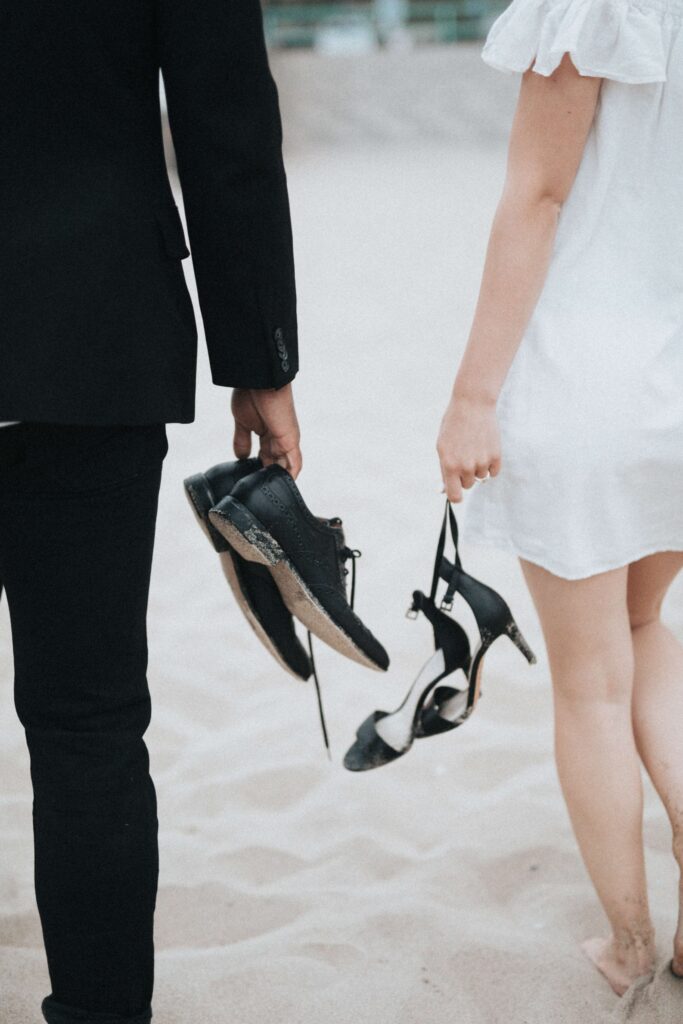A man and a woman wearing formal attire and holding formal shoes on a beach