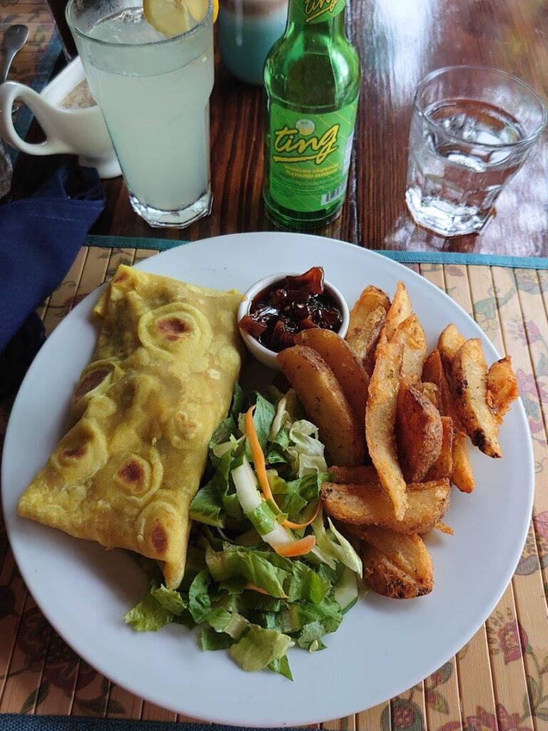 A traditional West Indian roti on a plate with some homemade chips and salad. next to the plate is a green glass bottle of ting and glass filled with the beverage