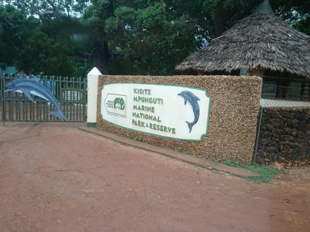 A stone wall with a white sign indicating Kisite Mpunguti Marine Park and Reserve with a blue dolphin image on a gate