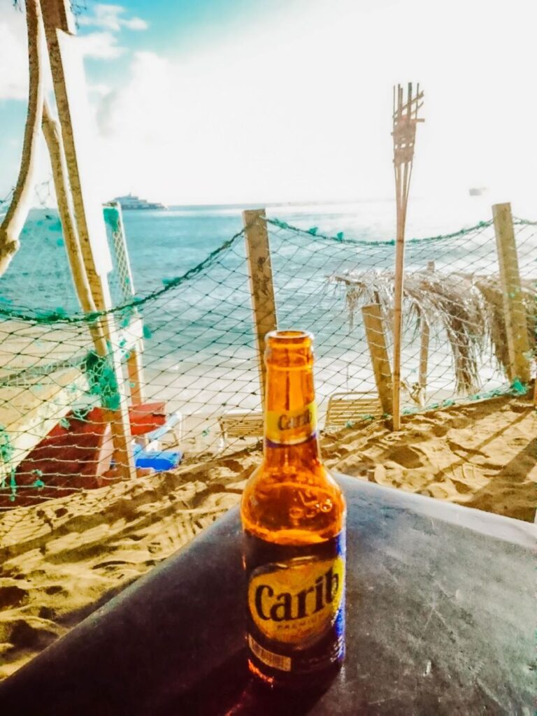 A brown beer bottle featuring the logo Carib sits on a black table in front of the sea 