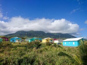 the forefront of the picture is green grass behind sits a group of colourful houses in St Kitts with a mountain in the background